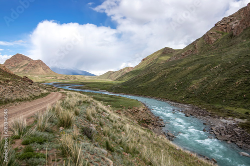View of the river flowing between green hills with grass and stones in the foreground Kyrgyzstan. Tien Shan