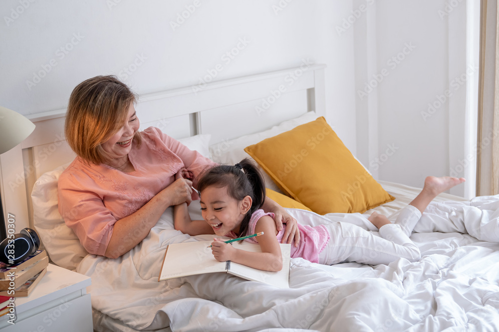 Happy little cute asian girl and her senior retired grandmother lying on the bed. Grandma smile and teach her preschool grandchild do homework or write on the book on the bed in bedroom. Happy little cute asian girl and her senior retired grandmother lying on the bed. Grandma smile and teach her preschool grandchild do homework or write on the book on the bed in bedroom.
