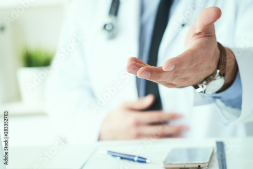 Male doctor making welcome gesture  politely inviting patient to sit down in medical office. Photo with depth of field.