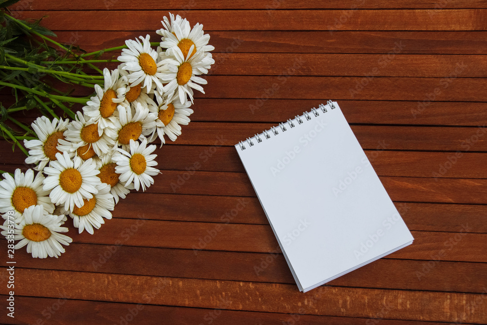 Obraz premium a bouquet of white daisies on a brown wooden table with a clean white notepad, copy space, flat lay, top view.