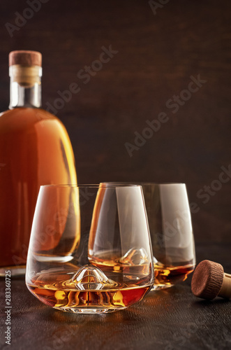 Two crystal glasses of whiskey and a full bottle on a wooden table, as well as on the table is a cork from the bottle.