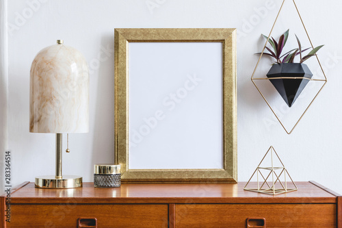 Stylish composition of open space interior with gold mock up frame,retro wooden commode, marble table lamp, gold pyramid, plants and elegant accessories. Modern design interior. Template. Ready to use photo