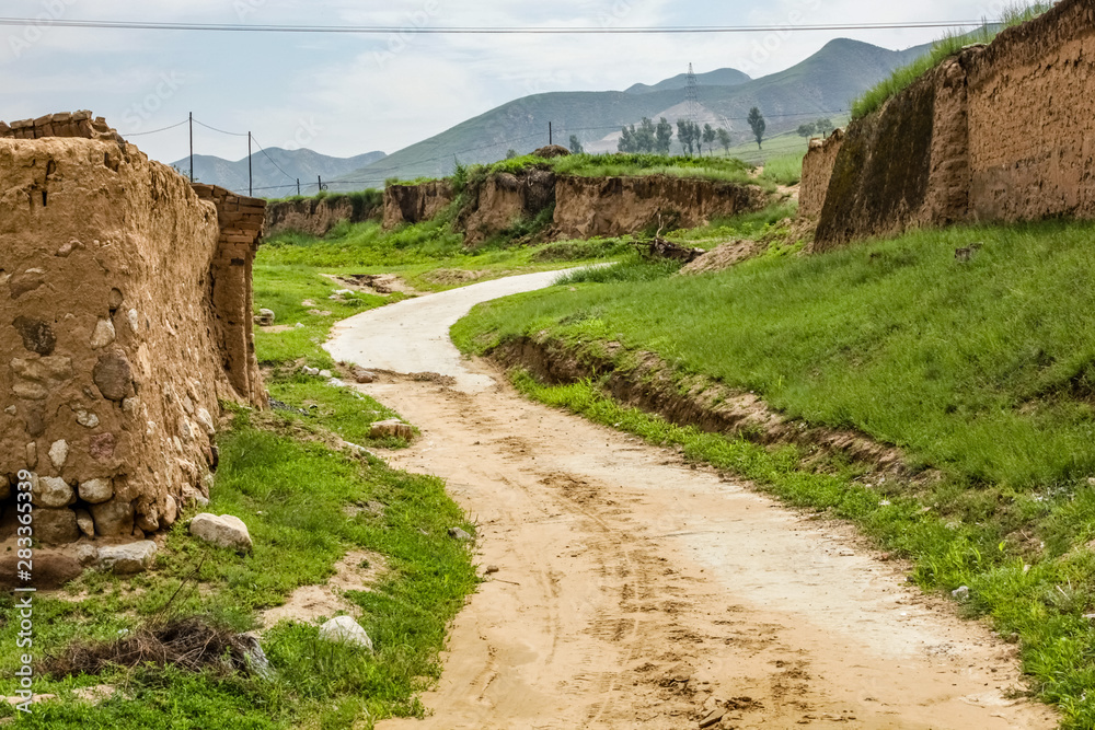 A dirt road winds up a small hill around a mud wall in rural China with a power line running across the top