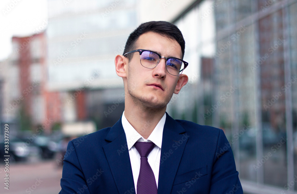 Serious self-confident businessman in formal suit and tie looking straight at camera in glasses outside. Handsome sad tired formal dressed guy standing outside office building or business centre. 