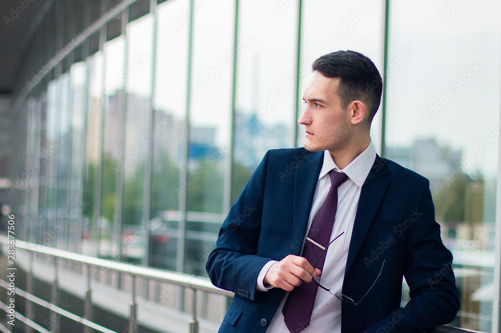 Pensive serious businessman, formal dressed thoughtful young guy in suit standing outside office modern building or business centre, holding glasses in hand, leans on railing outdoor. Confident male Pensive serious businessman, formal dressed thoughtful young guy in suit standing outside office modern building or business centre, holding glasses in hand, leans on railing outdoor. Confident male