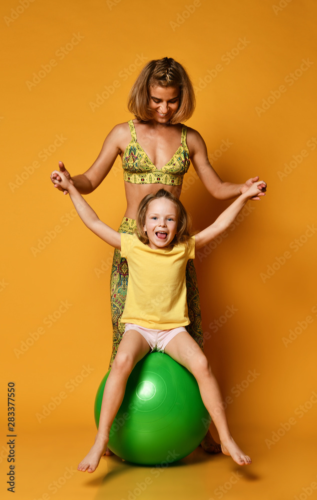 Cute child girl stretching on pilates fitness ball with mother isolated on yellow background.