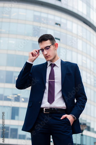 Handsome confident pensive businessman in a suit and tie holding his glasses and hand in a pocket, standing outdoor in front of office modern building, skyscraper or business center. Smart guy, male.