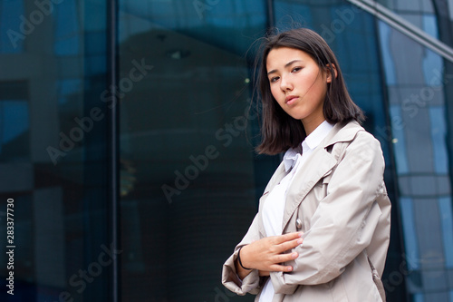 Serious young Asian student or Chinese businesswoman standing outside office workplace with textbooks or documents. Smart university female with books outdoor looking at camera in white shirt, cloak.  © Евгений Шемякин