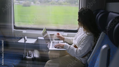 Young girl travelling on train in daytime. Side view charming woman sitting near window, working on computer. Female drinking coffee and enjoying landscape view passing by at background. 
