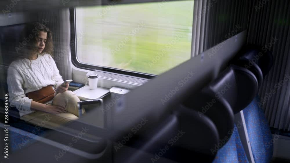 Young girl travelling on train in daytime. Medium shot woman sitting near window with landscape view passing by, view reflection in mirror female using cell phone for messaging and communication
