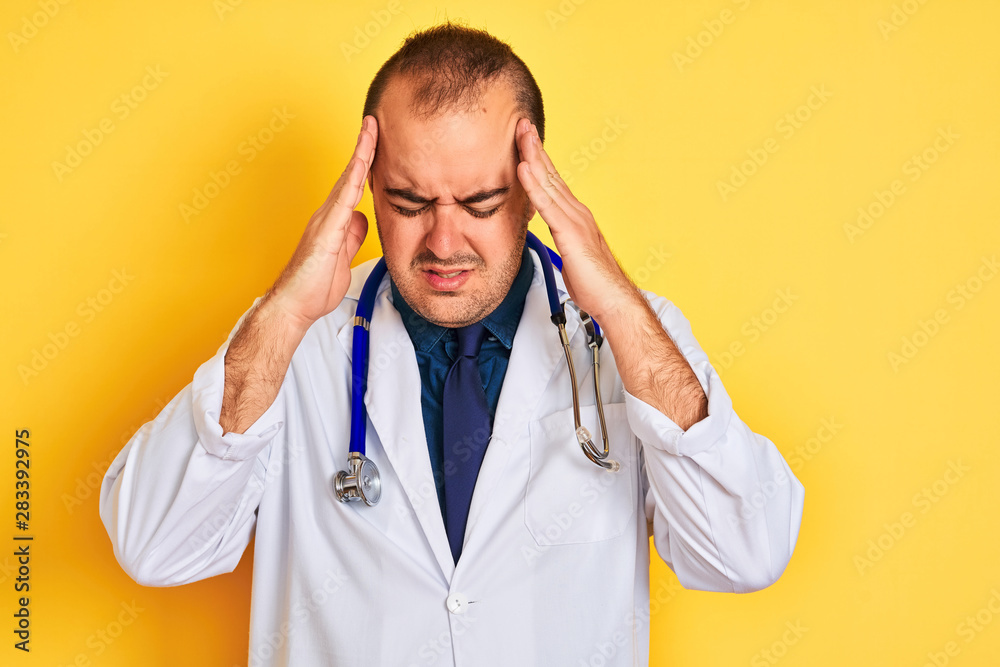 Young doctor man wearing coat and stethoscope standing over isolated yellow background with hand on head for pain in head because stress. Suffering migraine. Young doctor man wearing coat and stethoscope standing over isolated yellow background with hand on head for pain in head because stress. Suffering migraine.