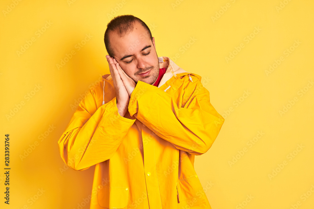 Young man wearing rain coat standing over isolated yellow background sleeping tired dreaming and posing with hands together while smiling with closed eyes.