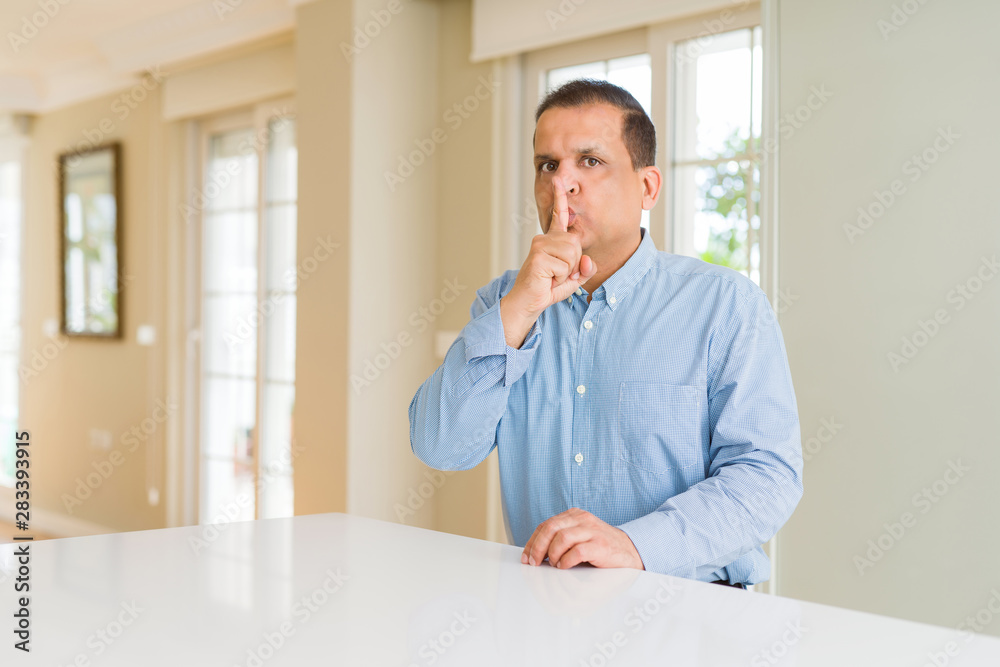 Middle age man sitting at home asking to be quiet with finger on lips. Silence and secret concept.