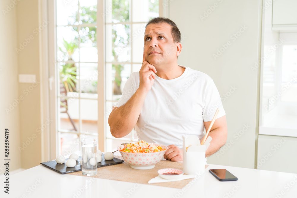 Middle age man eating asian food with chopsticks at home with hand on chin thinking about question, pensive expression. Smiling with thoughtful face. Doubt concept. Middle age man eating asian food with chopsticks at home with hand on chin thinking about question, pensive expression. Smiling with thoughtful face. Doubt concept.