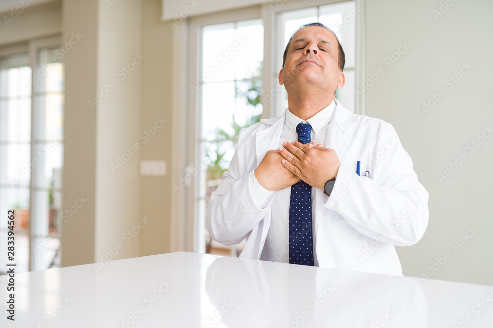 Middle age doctor man wearing medical coat at the clinic smiling with hands on chest with closed eyes and grateful gesture on face. Health concept.