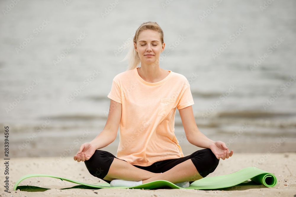 Fototapeta premium Pretty young girl relaxing siting in the lotus position on a yoga mat on sandy beach on a warm day