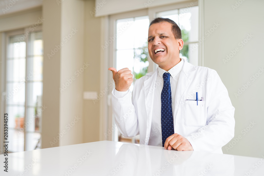 Middle age doctor man wearing medical coat at the clinic smiling with happy face looking and pointing to the side with thumb up.