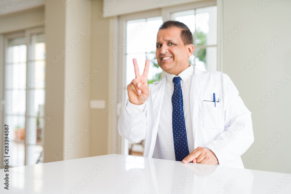 Middle age doctor man wearing medical coat at the clinic smiling looking to the camera showing fingers doing victory sign. Number two. Middle age doctor man wearing medical coat at the clinic smiling looking to the camera showing fingers doing victory sign. Number two.