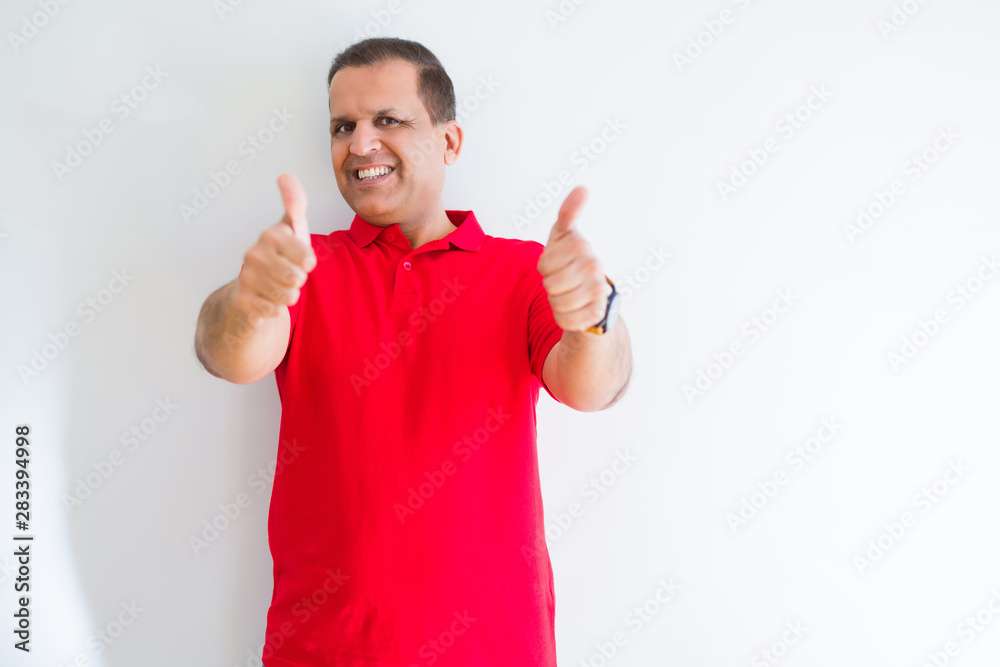 Middle age man wearing red t-shirt over white wall approving doing positive gesture with hand, thumbs up smiling and happy for success. Looking at the camera, winner gesture. Middle age man wearing red t-shirt over white wall approving doing positive gesture with hand, thumbs up smiling and happy for success. Looking at the camera, winner gesture.
