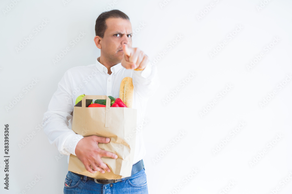 Middle age man holding groceries shopping bag over white background pointing with finger to the camera and to you, hand sign, positive and confident gesture from the front Middle age man holding groceries shopping bag over white background pointing with finger to the camera and to you, hand sign, positive and confident gesture from the front