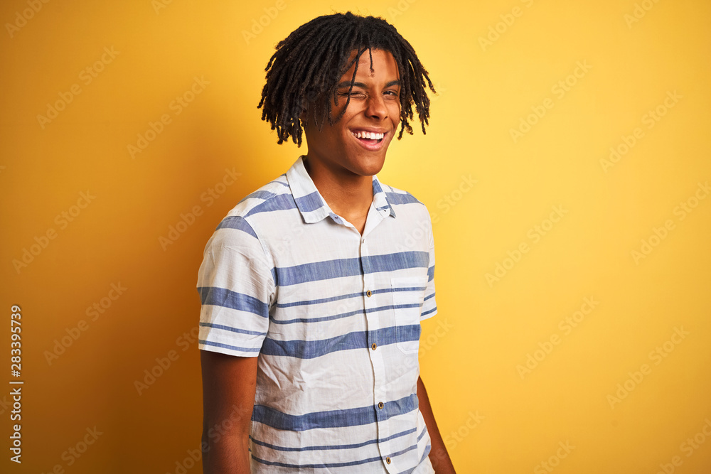 Afro american man with dreadlocks wearing striped shirt over isolated yellow background winking looking at the camera with sexy expression, cheerful and happy face. Afro american man with dreadlocks wearing striped shirt over isolated yellow background winking looking at the camera with sexy expression, cheerful and happy face.