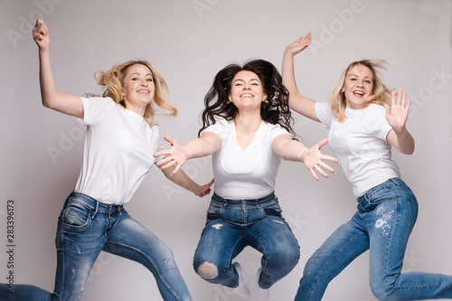 Close up of two blonde and one brunette women getting fun together. Three friends in casual clothes dancing and jumping. Beautiful happy girls in white t-shirts and jeans smiling and hanging out.