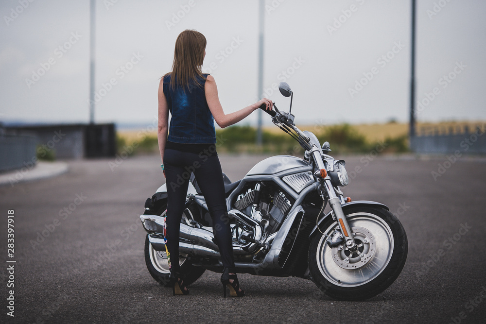 Fashion sexy woman is standing near classic style cruiser motorbike. Girl biker with perfect fit slim body on highway. The spirit of freedom and independence. Back view