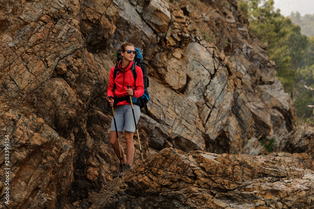 Smiling girl standing on the rocks with hiking backpack and walking sticks