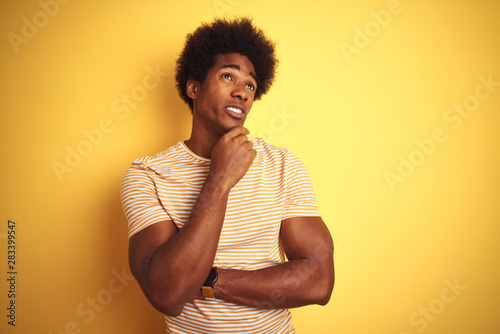 American man with afro hair wearing striped t-shirt standing over isolated yellow background Thinking worried about a question, concerned and nervous with hand on chin © Krakenimages.com