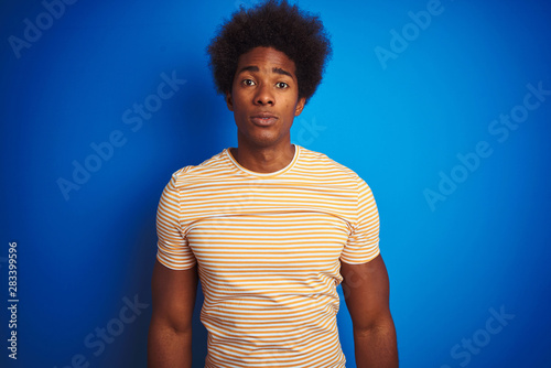 American man with afro hair wearing striped yellow t-shirt over isolated blue background with serious expression on face. Simple and natural looking at the camera.
