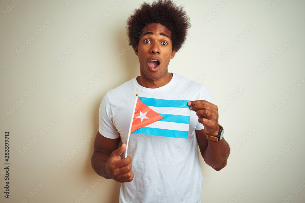 Young african american man holding Cuba Cuban flag standing over isolated white background scared in shock with a surprise face, afraid and excited with fear expression Young african american man holding Cuba Cuban flag standing over isolated white background scared in shock with a surprise face, afraid and excited with fear expression