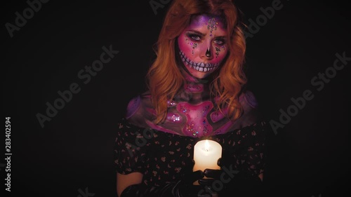 bright lady with color makeup in style of day of dead, woman with rhinestones on face in image of St. Katrina, sweet pink and violet shiny cheper with burning candle in hands with open shoulders photo