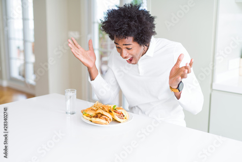 African American hungry man eating hamburger for lunch celebrating crazy and amazed for success with arms raised and open eyes screaming excited. Winner concept