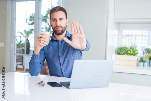 Handsome man working using computer laptop and drinking a cup of coffee with open hand doing stop sign with serious and confident expression, defense gesture