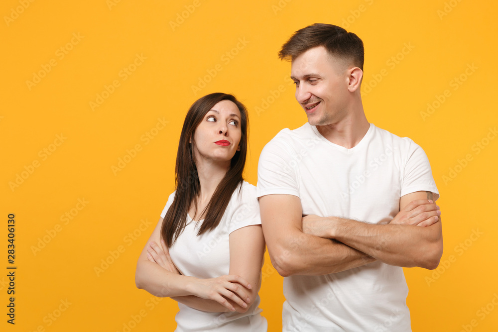Smirked young couple two friends guy girls in white blank empty design t-shirts posing isolated on yellow orange wall background. People lifestyle concept. Mock up copy space. Holding hands crossed. Smirked young couple two friends guy girls in white blank empty design t-shirts posing isolated on yellow orange wall background. People lifestyle concept. Mock up copy space. Holding hands crossed.