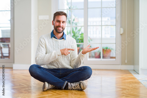 Handsome man wearing casual sweater sitting on the floor at home amazed and smiling to the camera while presenting with hand and pointing with finger.