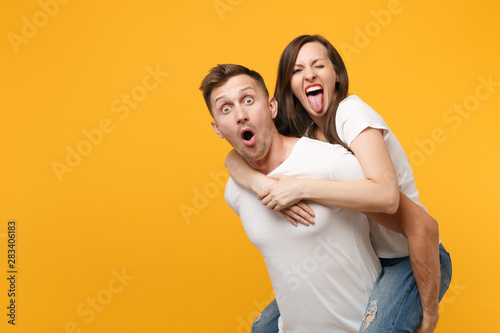 Shocked young couple two friends guy girl in white t-shirts posing isolated on yellow orange background. People lifestyle concept. Mock up copy space. Giving piggyback ride to joyful, sitting on back.