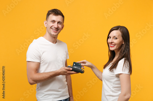 Young woman and man in white t-shirts posing isolated on yellow orange background. People lifestyle concept. Mock up copy space. Hold wireless payment terminal to process acquire credit card payments.
