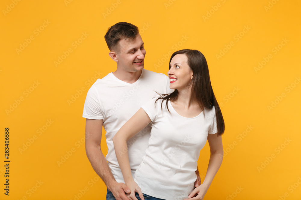Cheerful young couple two friends guy girl in white empty blank design t-shirts posing isolated on yellow orange wall background. People lifestyle concept. Mock up copy space. Looking at each other. Cheerful young couple two friends guy girl in white empty blank design t-shirts posing isolated on yellow orange wall background. People lifestyle concept. Mock up copy space. Looking at each other.