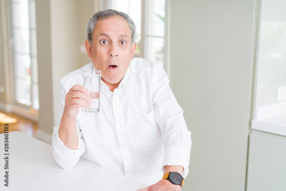Handsome senior man drinking a fresh glass of water at home scared in shock with a surprise face, afraid and excited with fear expression