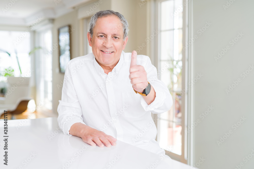 Handsome senior man at home doing happy thumbs up gesture with hand. Approving expression looking at the camera with showing success.