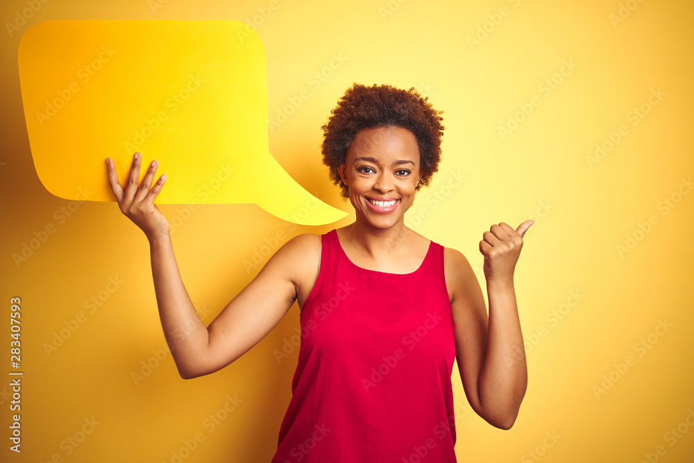 Young african american woman holding speech bubble over yellow isolated background pointing and showing with thumb up to the side with happy face smiling Young african american woman holding speech bubble over yellow isolated background pointing and showing with thumb up to the side with happy face smiling