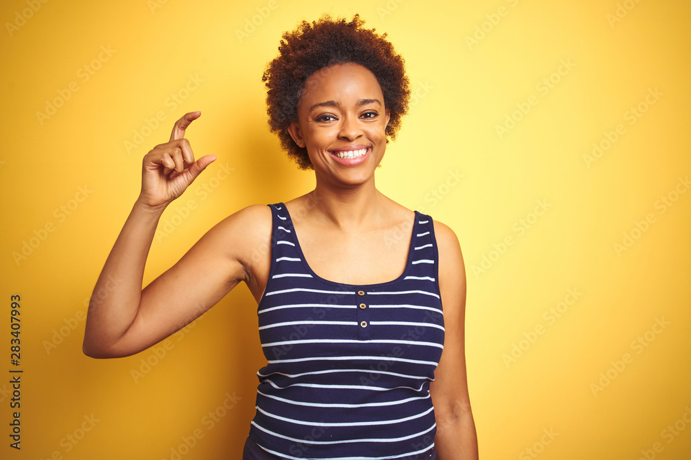 Beauitul african american woman wearing summer t-shirt over isolated yellow background smiling and confident gesturing with hand doing small size sign with fingers looking and the camera. 