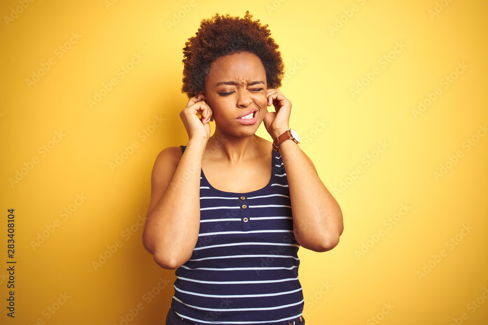 Beauitul african american woman wearing summer t-shirt over isolated yellow background covering ears with fingers with annoyed expression for the noise of loud music. Deaf concept. Beauitul african american woman wearing summer t-shirt over isolated yellow background covering ears with fingers with annoyed expression for the noise of loud music. Deaf concept.