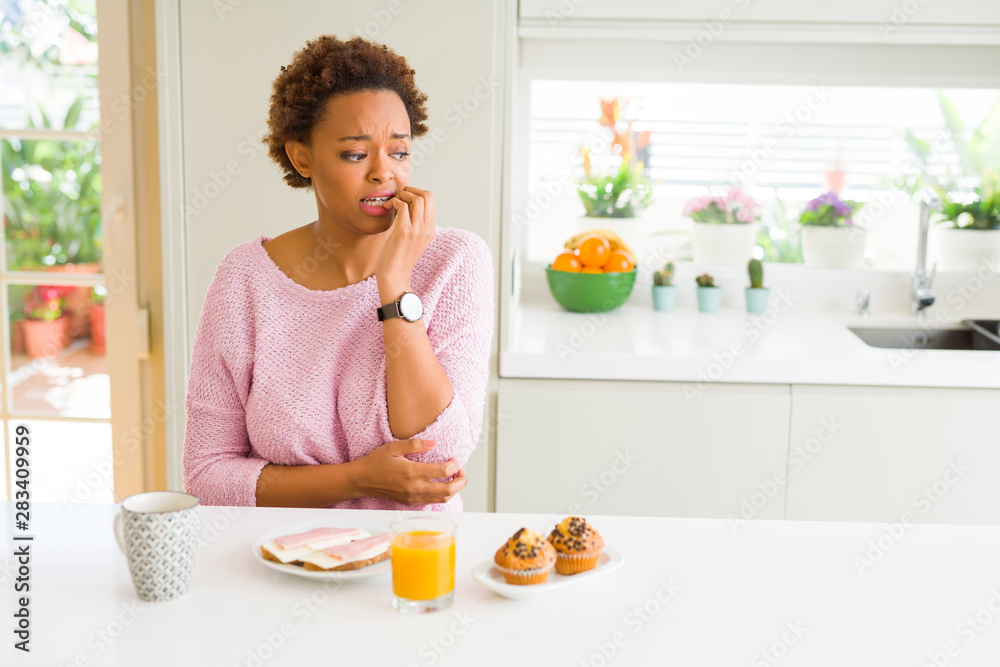 Young african american woman eating breaksfast in the morning at home looking stressed and nervous with hands on mouth biting nails. Anxiety problem.