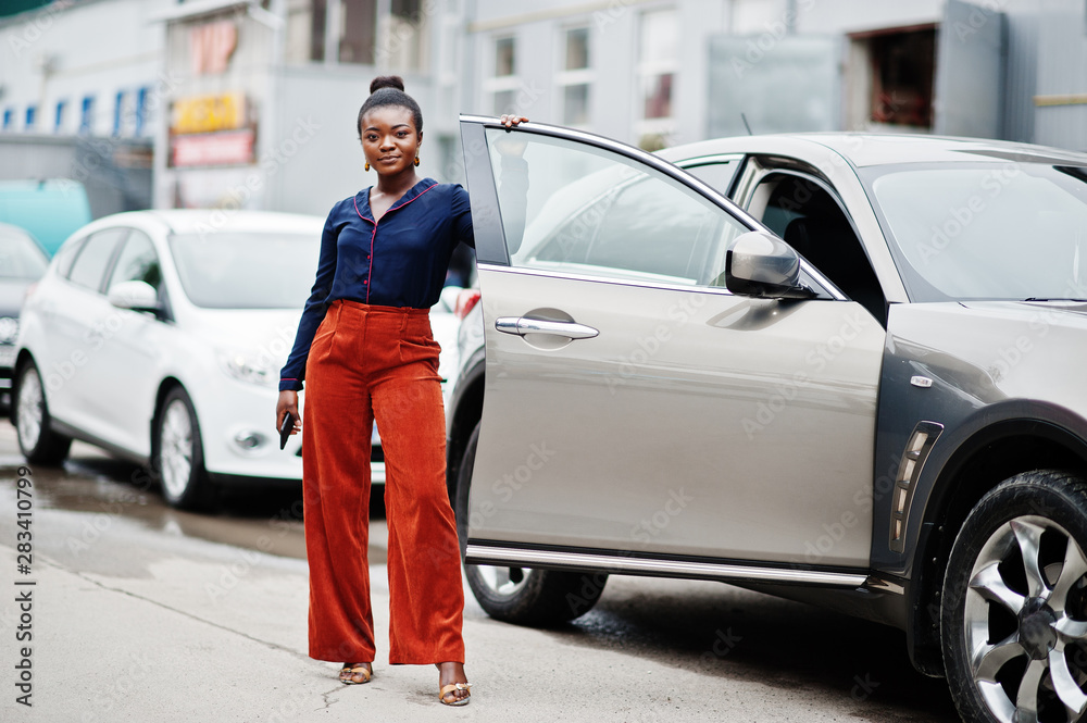 Rich business african woman in orange pants and blue shirt posed against silver suv car with opened door.