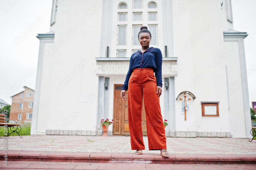 African woman in orange pants and blue shirt posed against large church. Faith and believe on God.