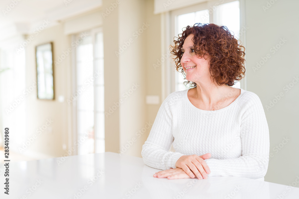 Beautiful senior woman wearing white sweater at home looking away to side with smile on face, natural expression. Laughing confident. Beautiful senior woman wearing white sweater at home looking away to side with smile on face, natural expression. Laughing confident.