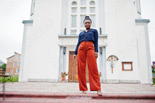 African woman in orange pants and blue shirt posed against large church. Faith and believe on God. © AS Photo Family