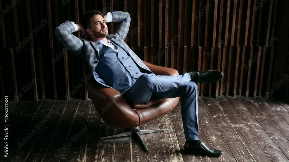 Young handsome businessman with a beard in a chic blue suit is resting. the lawyer is sitting in a leather chair by the window. Young handsome businessman with a beard in a chic blue suit is resting. the lawyer is sitting in a leather chair by the window.